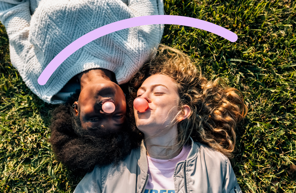 Two people lie on green grass, blowing pink bubblegum